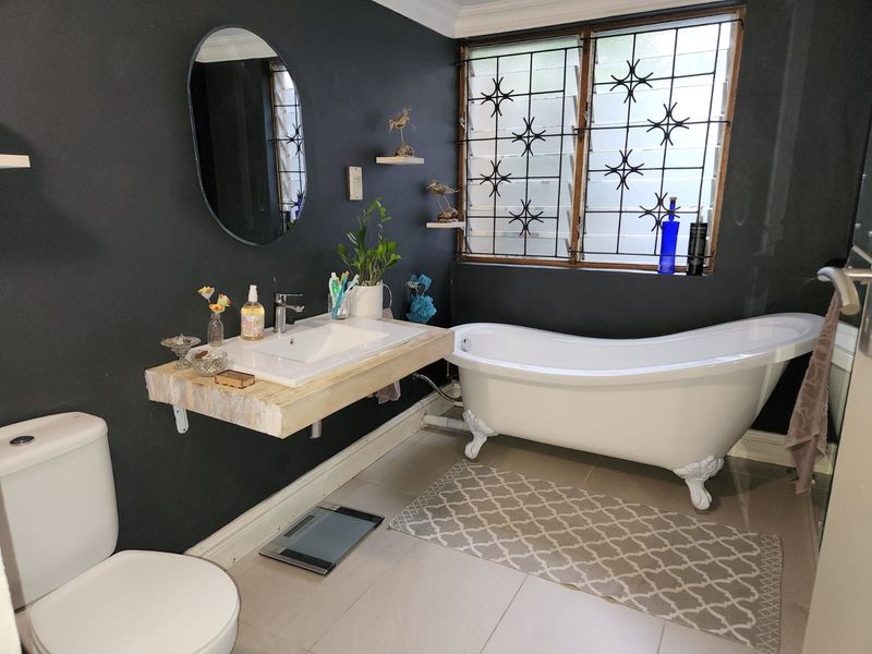 A bathroom featuring a freestanding clawfoot bathtub, a wall-mounted vanity with a ceramic basin, and dark painted walls with security-barred windows.