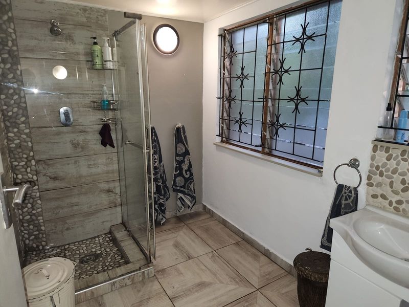 A functional bathroom featuring a glass-enclosed shower with pebble flooring, wood-look wall tiles, and a window with security bars.