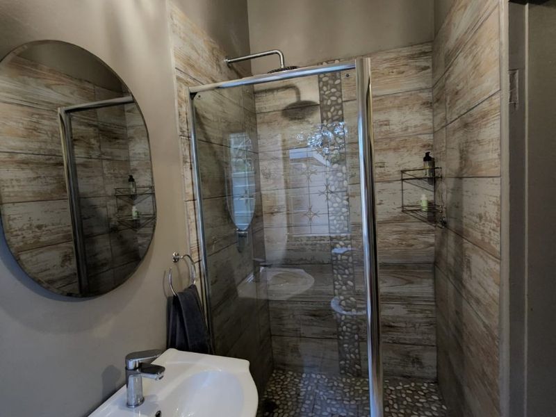 Modern bathroom featuring a vanity with basin, a glass-enclosed shower with pebble flooring, and wood-look wall tiles, accented by a decorative crystal chandelier.