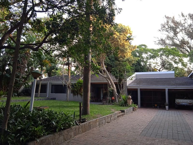 Front view of the property featuring a paved driveway, covered garage, and a well-established garden with mature trees and brick retaining walls.