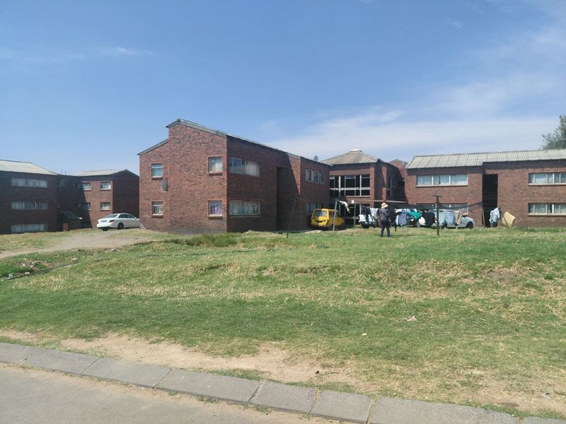 Exterior view of a multi-unit brick residential complex featuring a shared lawn area and corrugated metal roofing.