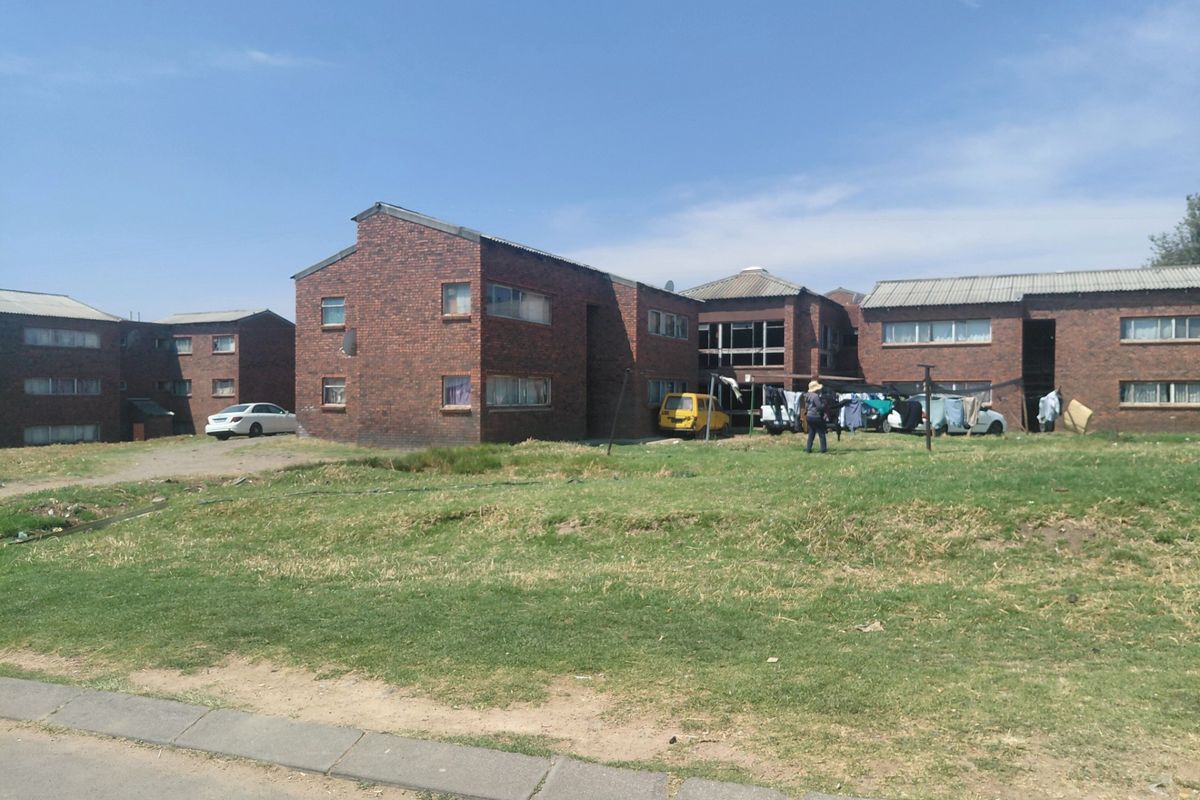 Exterior view of a multi-unit brick residential complex featuring a shared lawn area and corrugated metal roofing.