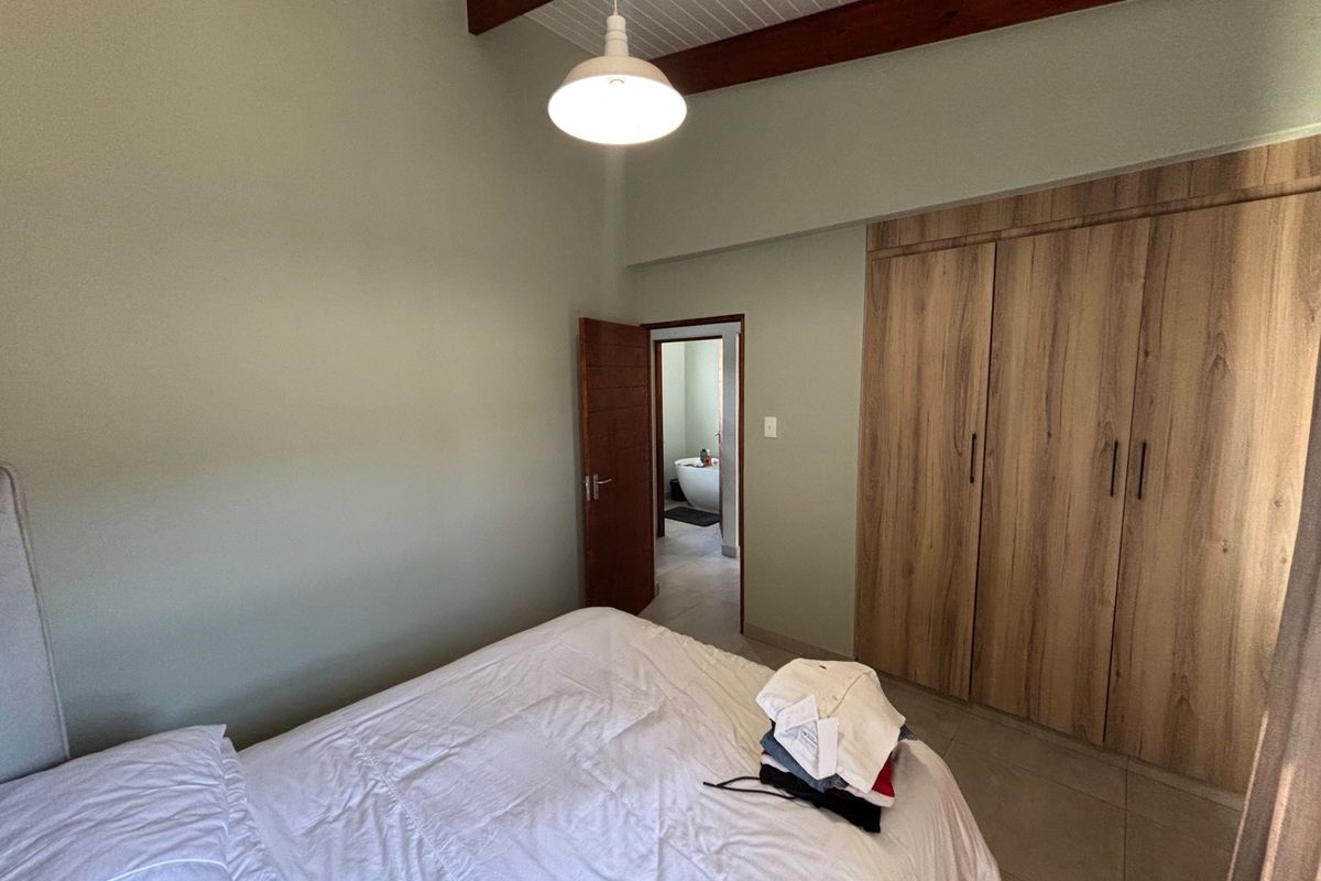A bedroom featuring built-in wooden cupboards, exposed timber ceiling beams, and tiled flooring, with an adjoining en-suite bathroom visible through the doorway.