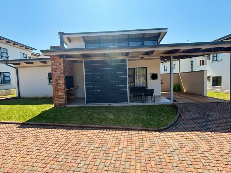 Front view of a modern townhouse featuring a paved driveway, carport, covered patio with a built-in braai, and distinctive clerestory windows.