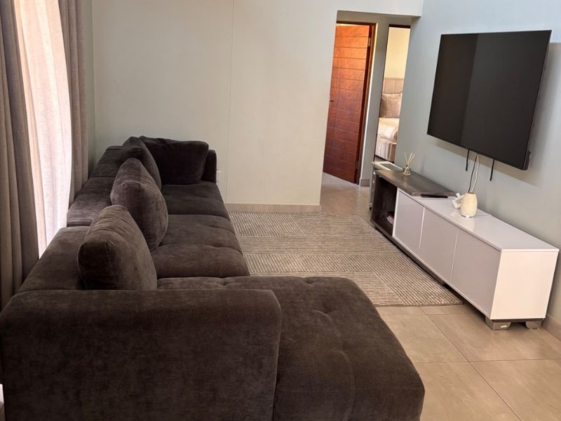 A bright living room featuring high vaulted ceilings, neutral tiled flooring, and a clean, modern layout with direct access to an adjoining bedroom.