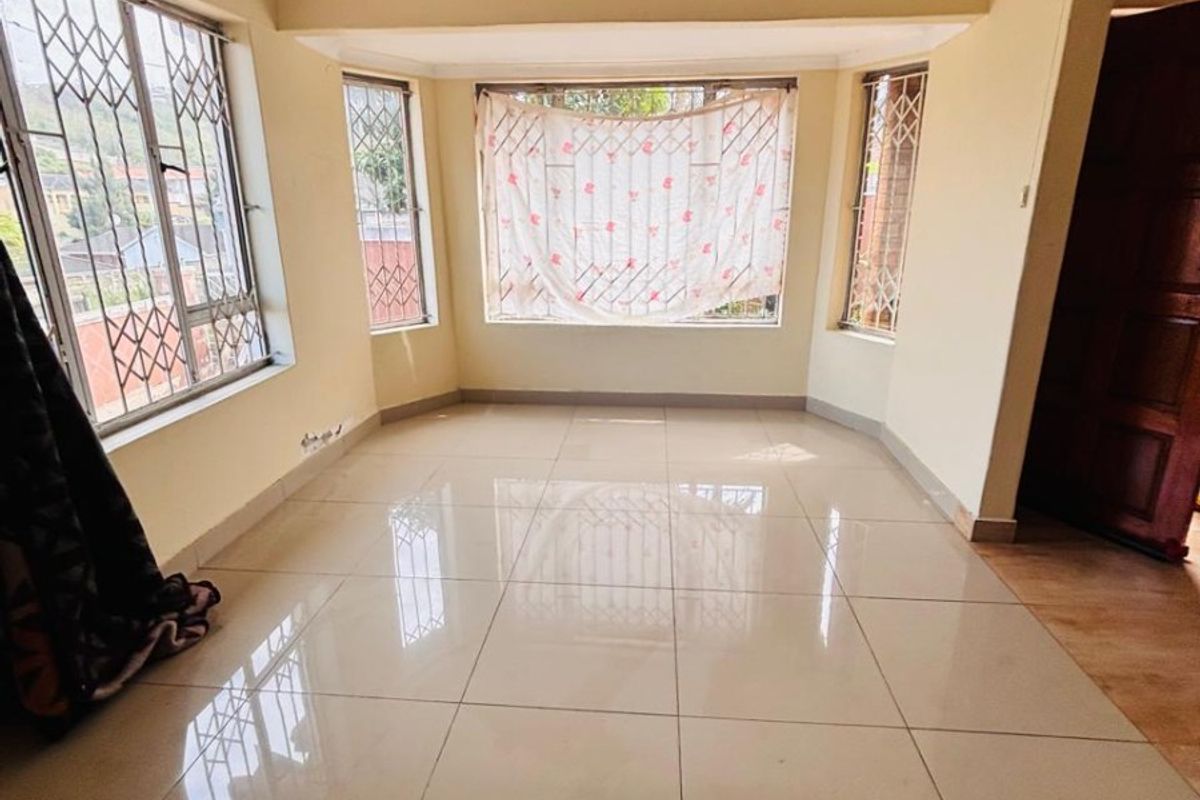 A bright, empty living room featuring large bay windows with security bars and neutral tiled flooring throughout.