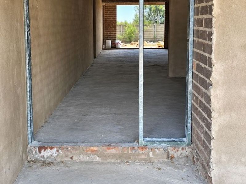 An unfinished hallway area under construction, featuring exposed brick walls, concrete flooring, and visible roof trusses, awaiting final interior finishes.