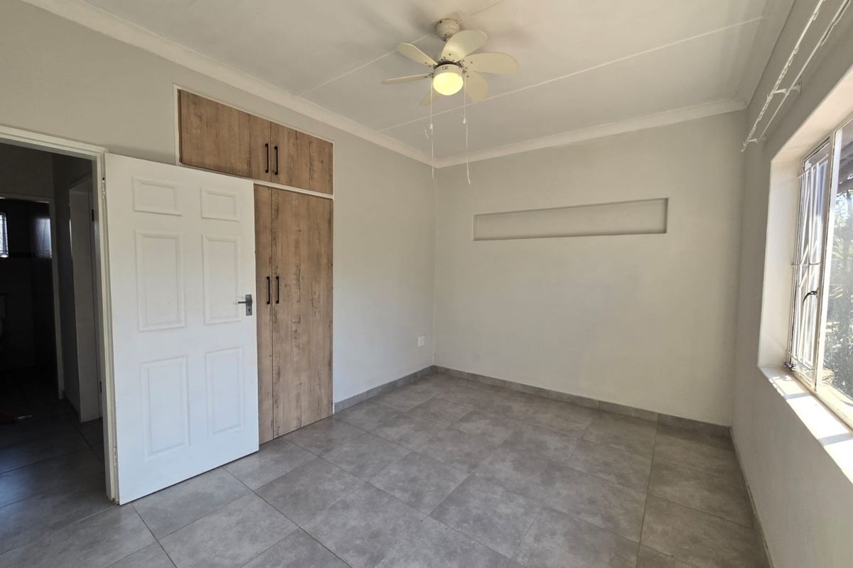 A bright, empty bedroom featuring neutral tiled flooring, built-in wood-finish cupboards, a ceiling fan, and a recessed wall niche.