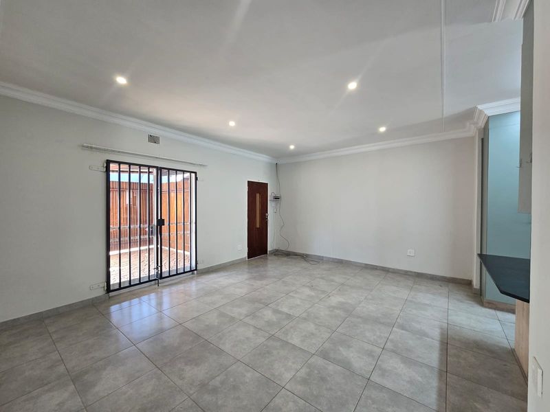 Spacious open-plan living area featuring neutral tiled flooring, recessed downlighting, decorative cornicing, and direct access to an outdoor area via a security gate.