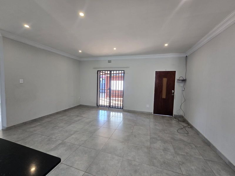 A spacious, empty living area featuring neutral tiled flooring, recessed downlights, and direct access to the exterior via a secure gate.