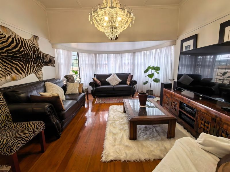 A well-maintained living room featuring polished hardwood flooring, a large bay window with natural light, and an elegant crystal chandelier.