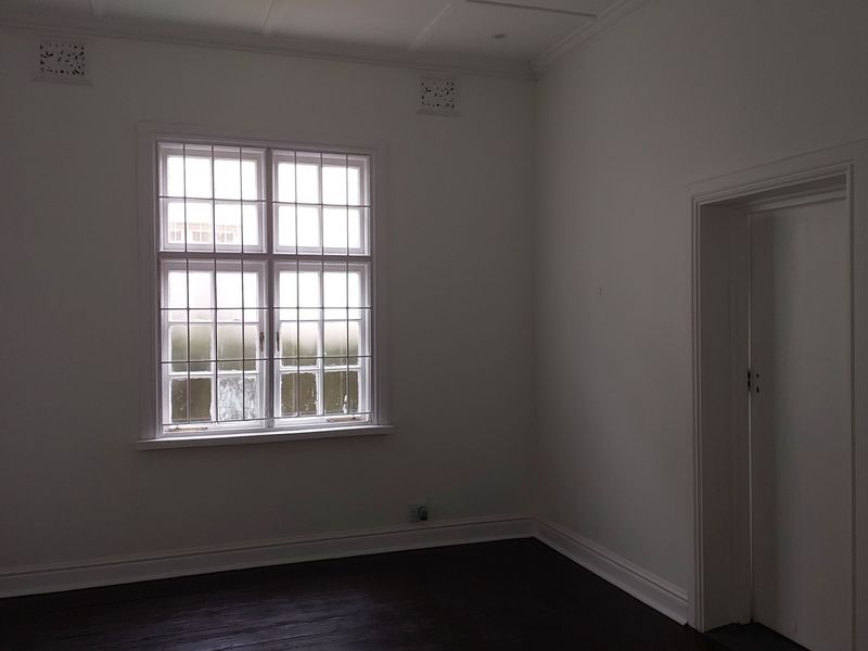 A bright, empty office space featuring dark timber flooring, white walls, and a window fitted with security bars.