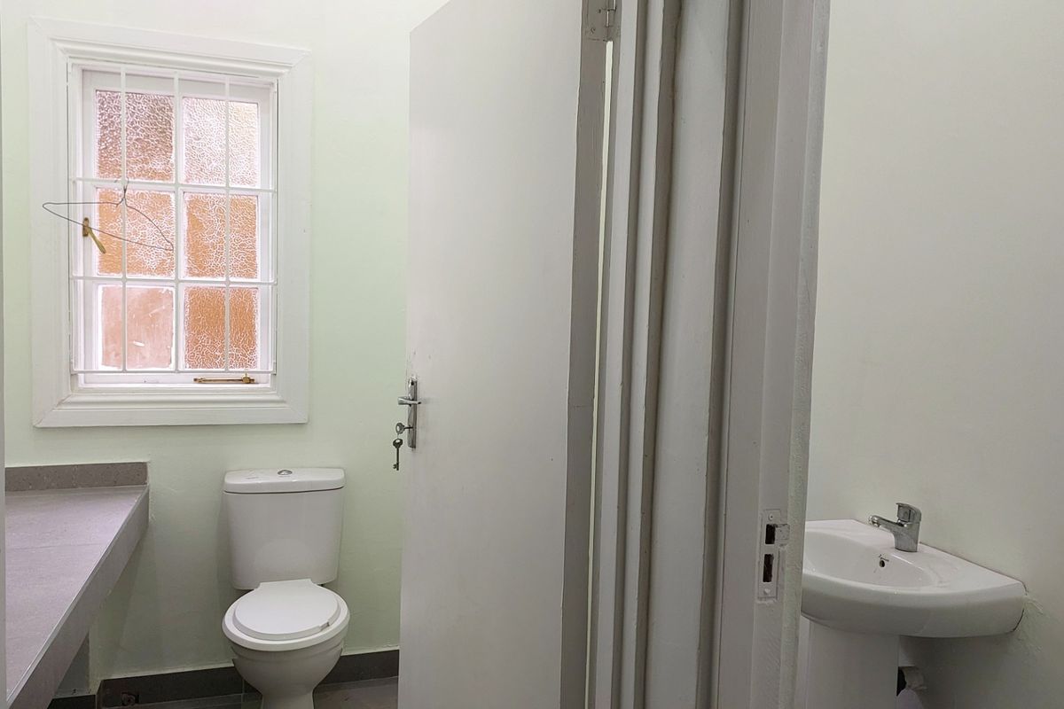 A functional bathroom featuring a pedestal basin, a toilet, and a built-in vanity counter, illuminated by a window with security bars.