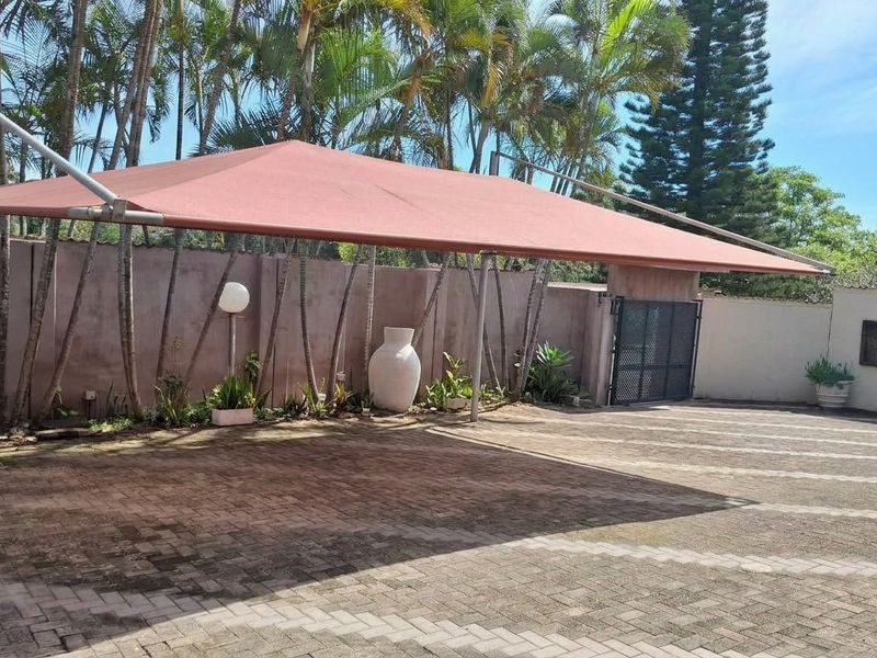 A spacious paved driveway area featuring a large shade sail structure, enclosed by a boundary wall and secure metal gate.