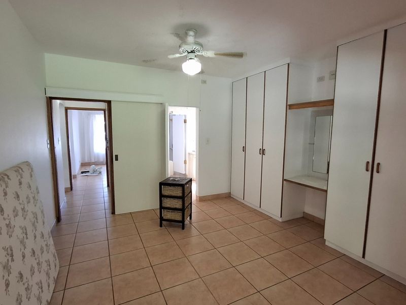 A spacious bedroom featuring tiled flooring, extensive built-in cupboards with a vanity section, and a ceiling fan, offering a functional layout.
