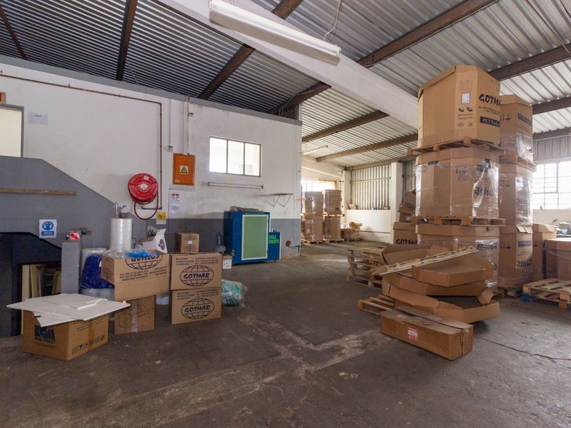 Industrial warehouse space featuring exposed steel roof trusses, corrugated metal roofing, concrete flooring, and essential fire safety equipment.