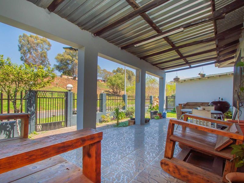 A covered outdoor patio area featuring tiled flooring, concrete support pillars, and a corrugated metal roof, overlooking the front garden and perimeter fencing.