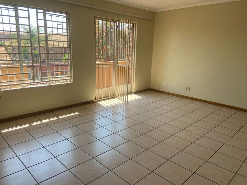 An empty, sunlit living area featuring neutral tiled flooring, security bars on windows, and a security gate leading to an outdoor space.