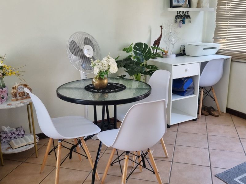 A living area featuring tiled flooring and venetian blinds, currently utilized as a multi-functional space with dining and desk areas.