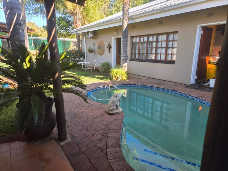 A backyard pool area featuring brick paving, a timber pergola, and direct access to the house with wooden window frames.