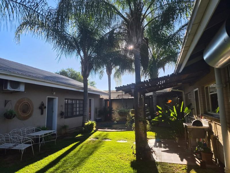 A well-maintained rear garden area featuring a lawn, mature palm trees, a covered patio with a pergola, and an external utility wash basin.