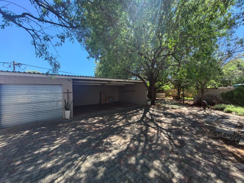 A spacious garage and carport area featuring a roll-up door and paved driveway, set within a mature, tree-lined garden environment.