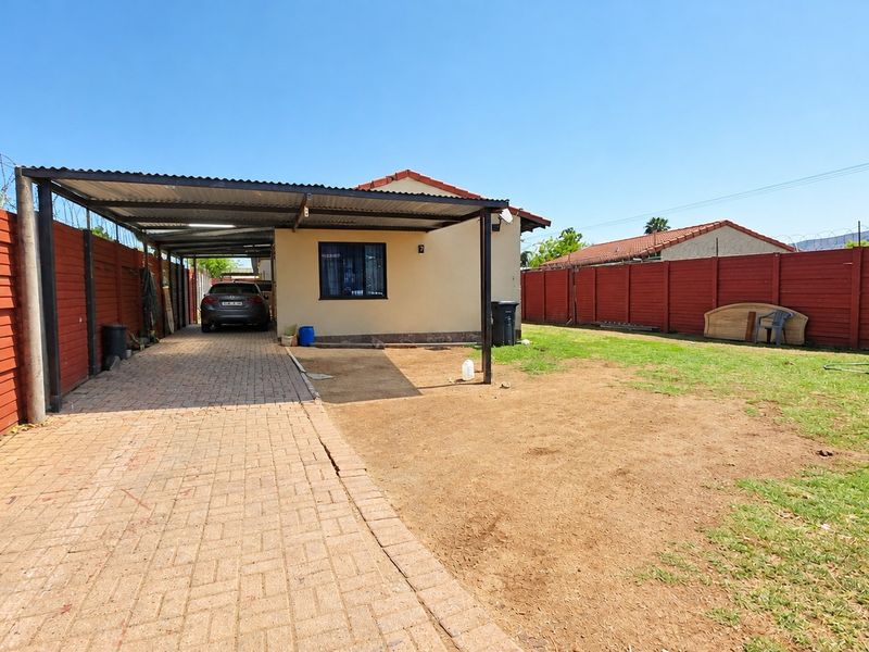 Rear exterior view featuring a paved driveway, a covered carport, and a fenced garden area with lawn.