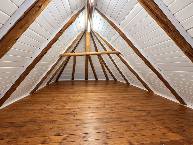 An attic-style bedroom featuring exposed timber rafters, polished wooden floorboards, and white tongue and groove ceiling paneling, offering a cozy, rustic aesthetic.