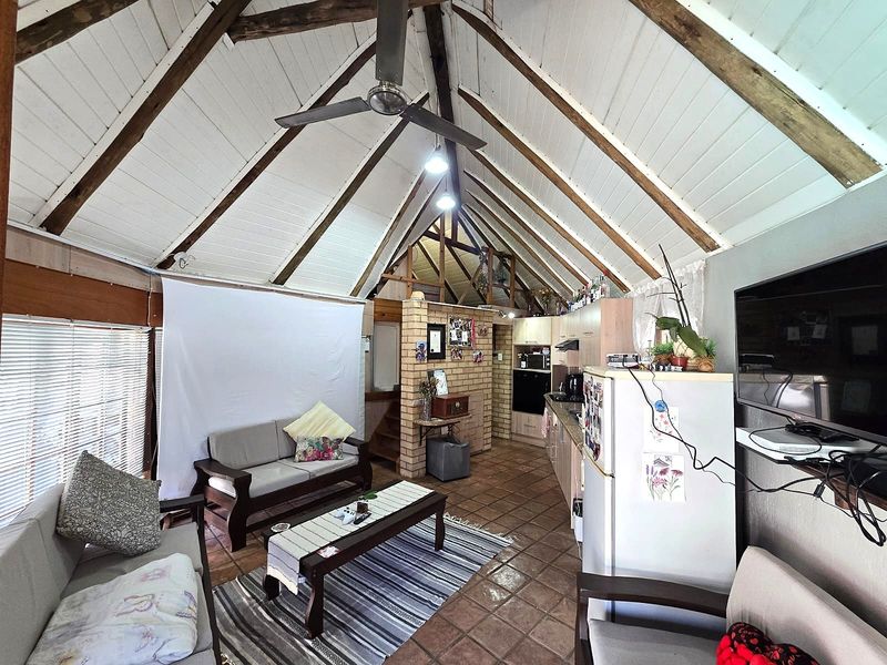 Open-plan living area featuring a vaulted ceiling with exposed timber rafters, tiled flooring, a brick fireplace, and an integrated kitchen space.