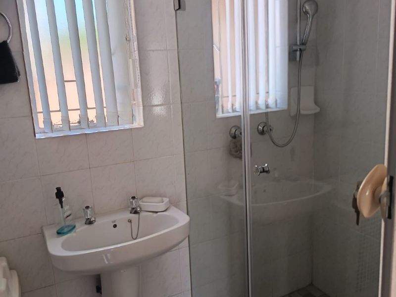 A functional bathroom featuring a pedestal washbasin, a glass-enclosed shower with a handheld fixture, and neutral wall tiling.