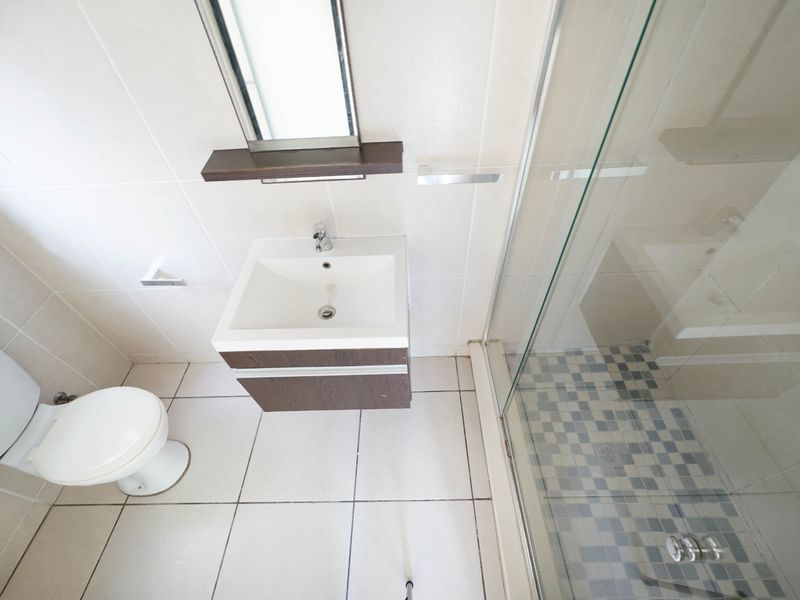 A clean, modern bathroom featuring a vanity basin, wall-mounted mirror, toilet, and a glass-enclosed shower with tiled finishes.