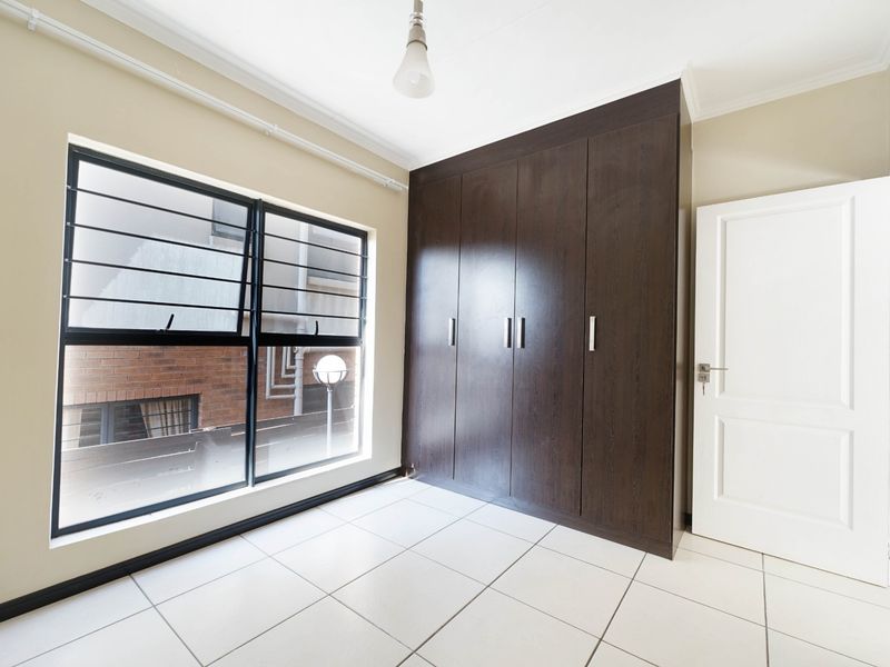 A bright, empty bedroom featuring dark wood built-in cupboards, tiled flooring, and a window fitted with security bars.