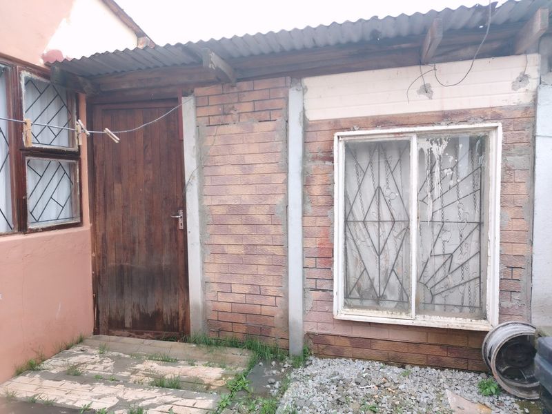 Rear exterior view of a brick structure featuring a corrugated iron roof, timber door, and window with security bars, showing signs of significant weathering.