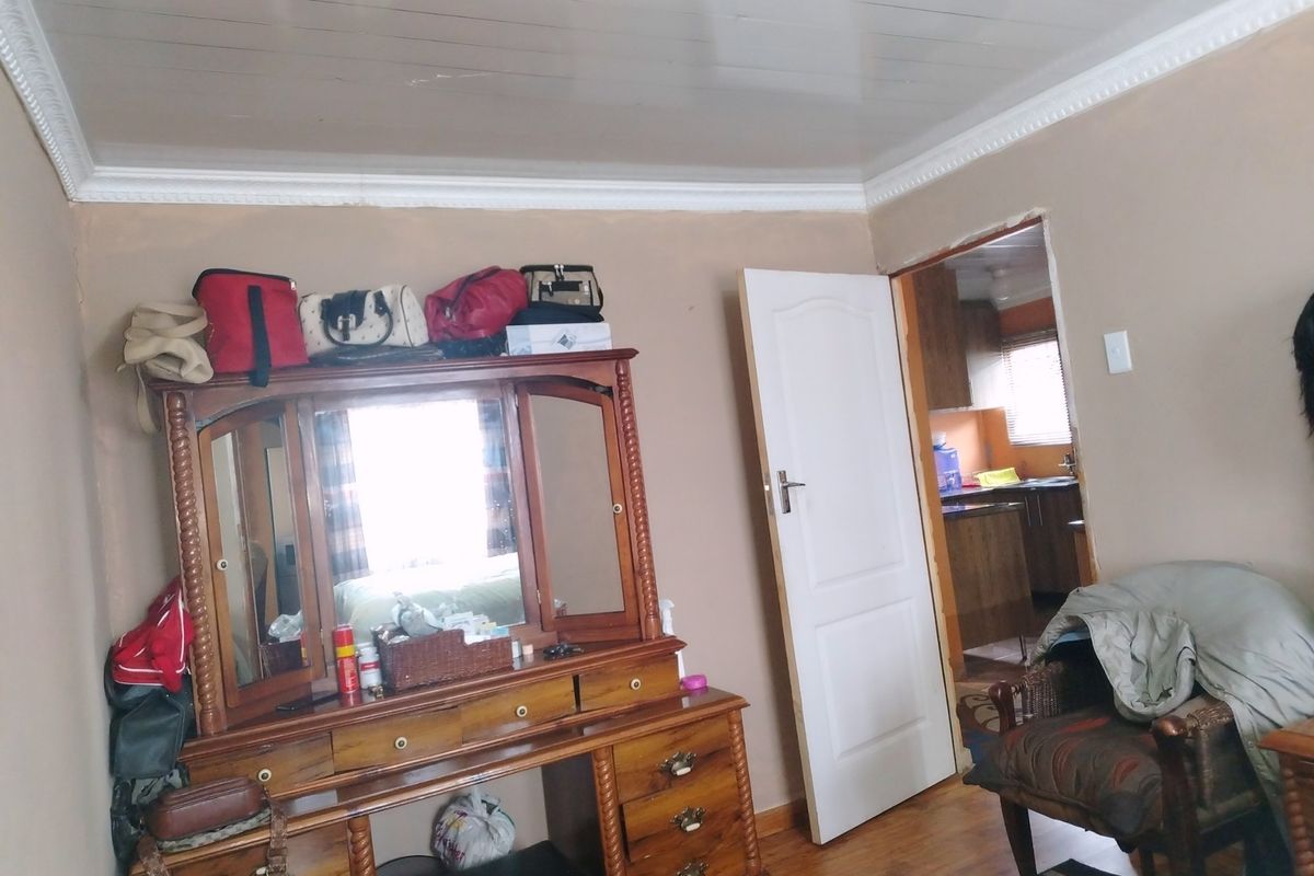 A bedroom featuring timber laminate flooring and coved ceiling cornices, with an adjacent view into the kitchen area.