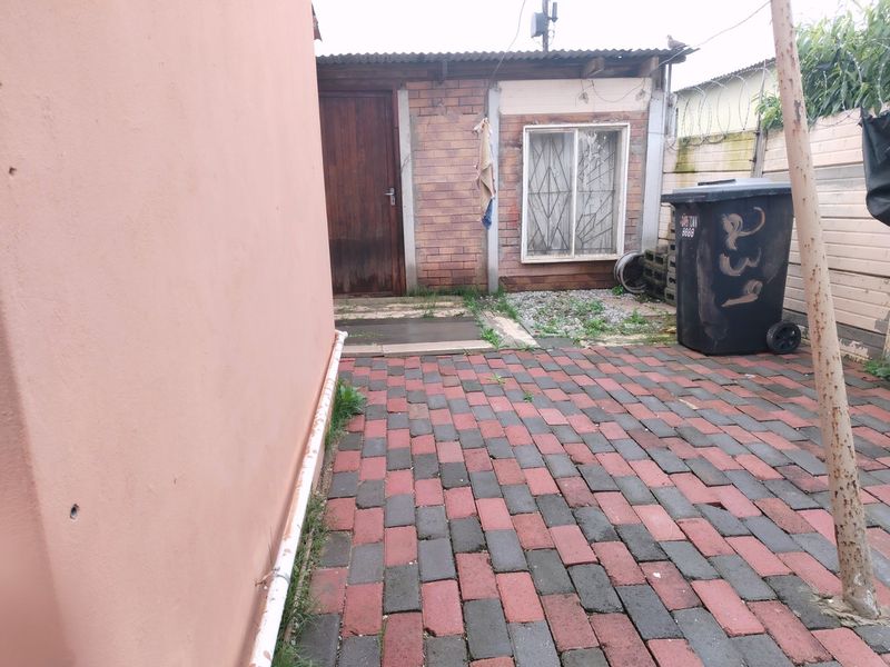 A rear exterior view showing a paved courtyard area with brick walling, a security-barred window, and a wooden door.