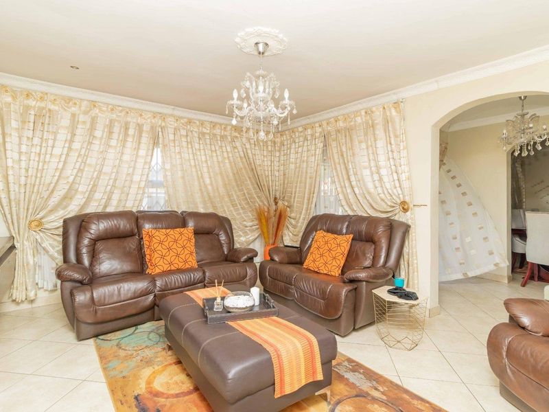 A well-maintained living room featuring decorative cornicing, a central ceiling rose with a crystal chandelier, and tiled flooring, leading into an adjacent dining area.