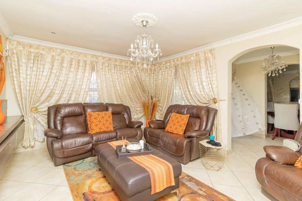 A well-maintained living room featuring decorative cornicing, a central ceiling rose with a crystal chandelier, and tiled flooring, leading into an adjacent dining area.