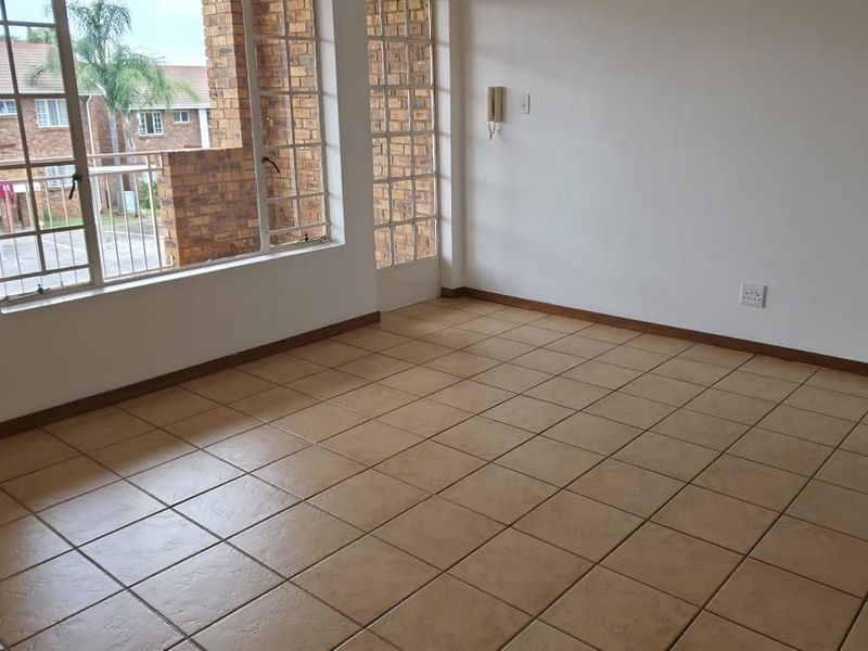 A bright, empty living area featuring neutral tiled flooring, an intercom system, and large windows providing natural light and views of the complex.