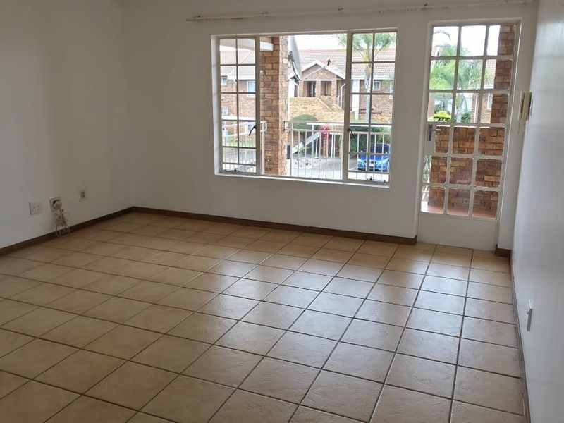 A bright, empty living room featuring neutral tiled flooring, large windows, and a glass panel door leading to an outdoor area.