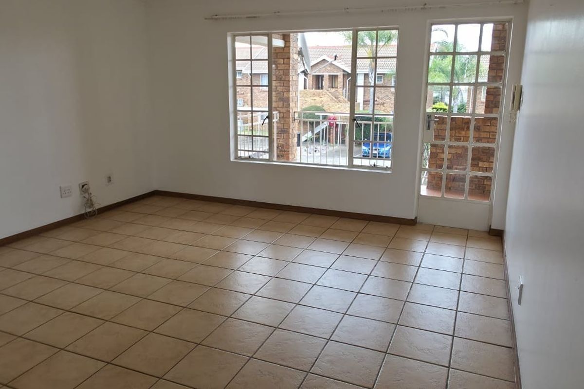A bright, empty living room featuring neutral tiled flooring, large windows, and a glass panel door leading to an outdoor area.