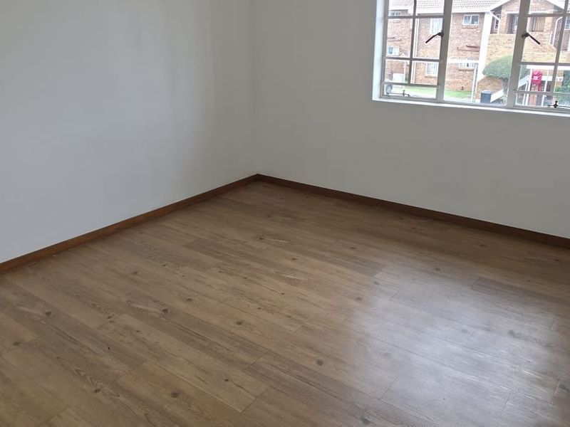 A bright, empty bedroom featuring modern laminate wood-look flooring, white walls, and a window with security bars.