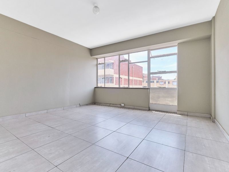A bright, empty living area featuring neutral wall tones, large windows providing natural light, and clean tiled flooring throughout.