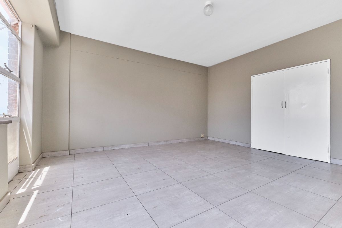A bright, empty bedroom featuring neutral wall tones, large windows for natural light, tiled flooring, and a built-in wardrobe.