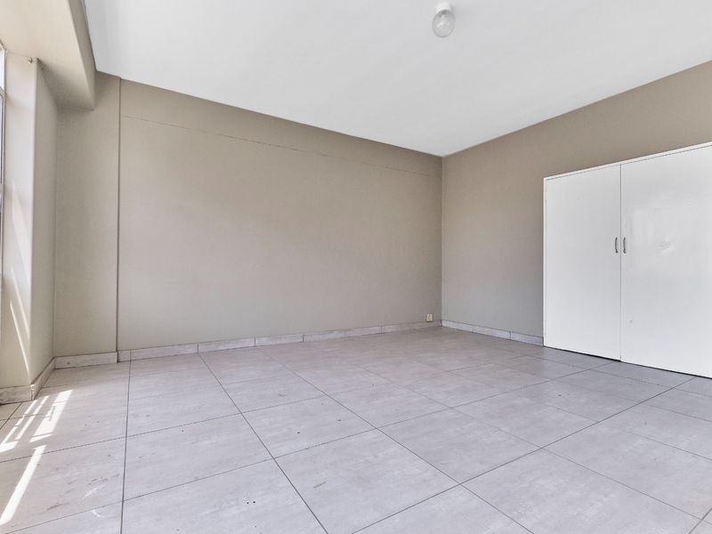 A bright, empty bedroom featuring neutral wall tones, large windows for natural light, tiled flooring, and a built-in wardrobe.