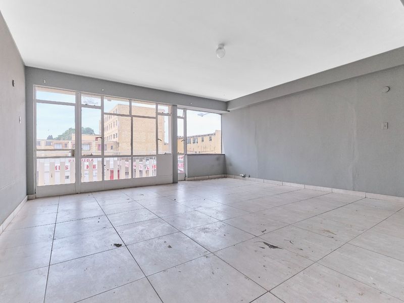 A spacious, empty living area featuring large windows, neutral grey walls, and tiled flooring, offering a blank canvas for renovation and interior design.