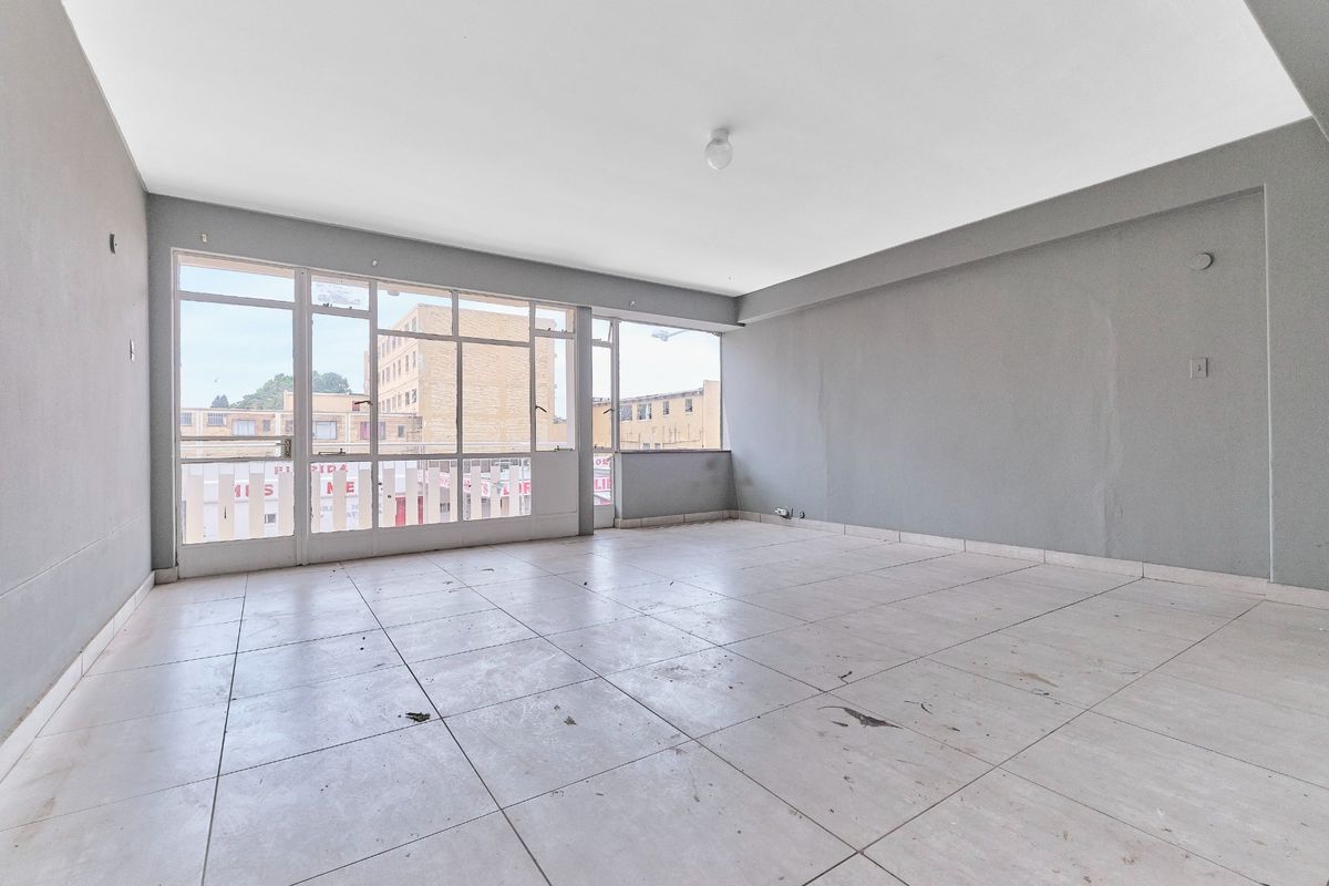 A spacious, empty living area featuring large windows, neutral grey walls, and tiled flooring, offering a blank canvas for renovation and interior design.