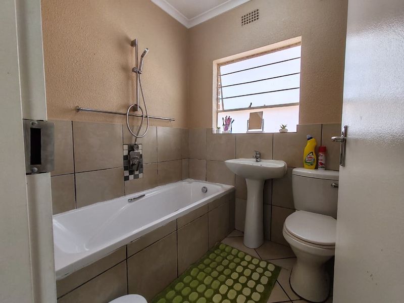 A functional bathroom featuring a bathtub with shower mixer, pedestal basin, and toilet, finished with neutral wall tiles and louver window.