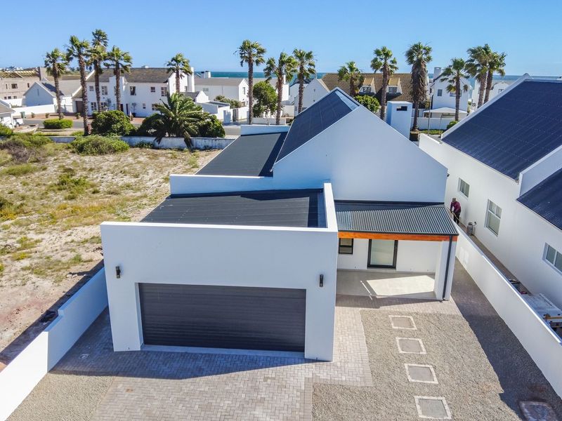 Aerial view of a modern white house featuring a garage, paved driveway, and a contemporary roof design in a coastal residential setting.