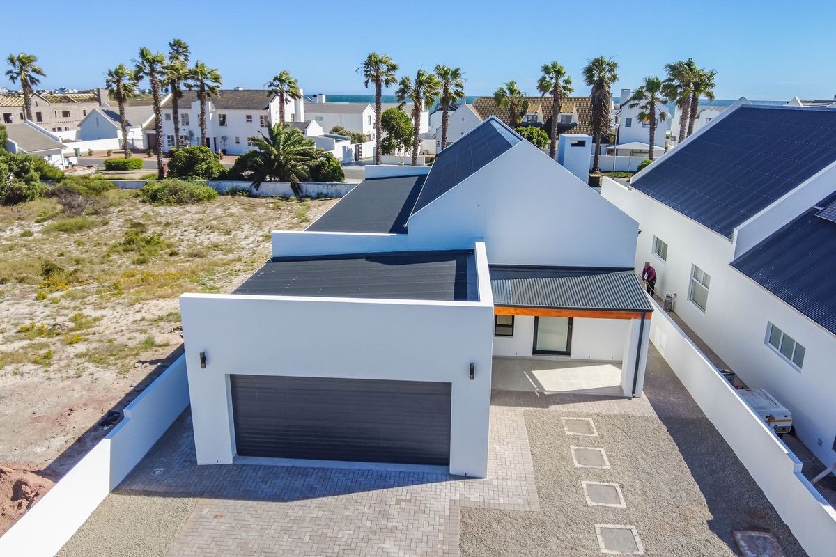 Aerial view of a modern white house featuring a garage, paved driveway, and a contemporary roof design in a coastal residential setting.