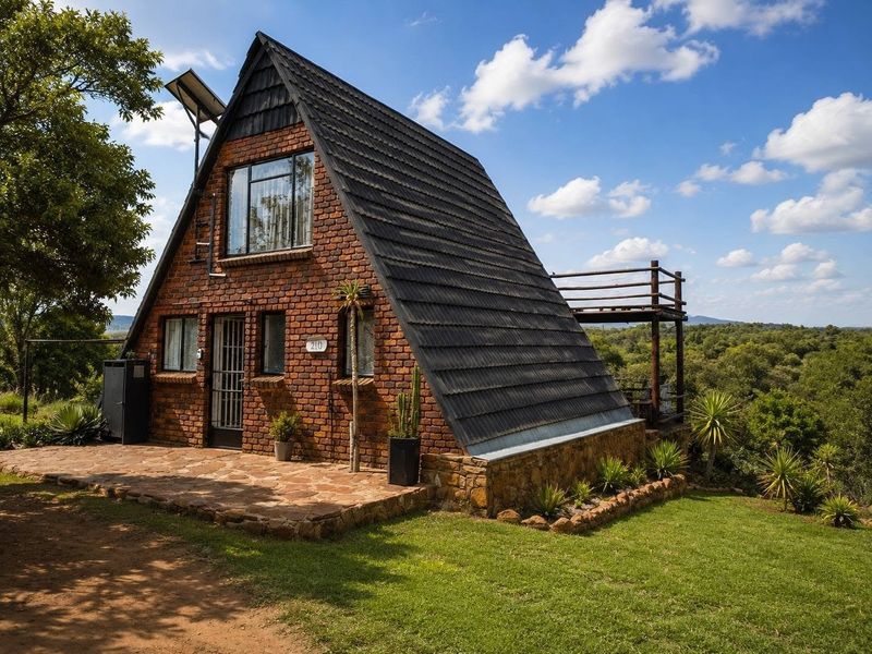 A charming A-frame brick house featuring a stone patio, raised wooden deck, and solar panel installation, set in a scenic natural environment.